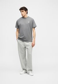 Νέος άνδρας που στέκεται με τα χέρια στις τσέπες, φορώντας γκρι μπλούζα New Balance, ανοιχτό γκρι φόρμα και λευκά αθλητικά παπούτσια σε απλό φόντο.