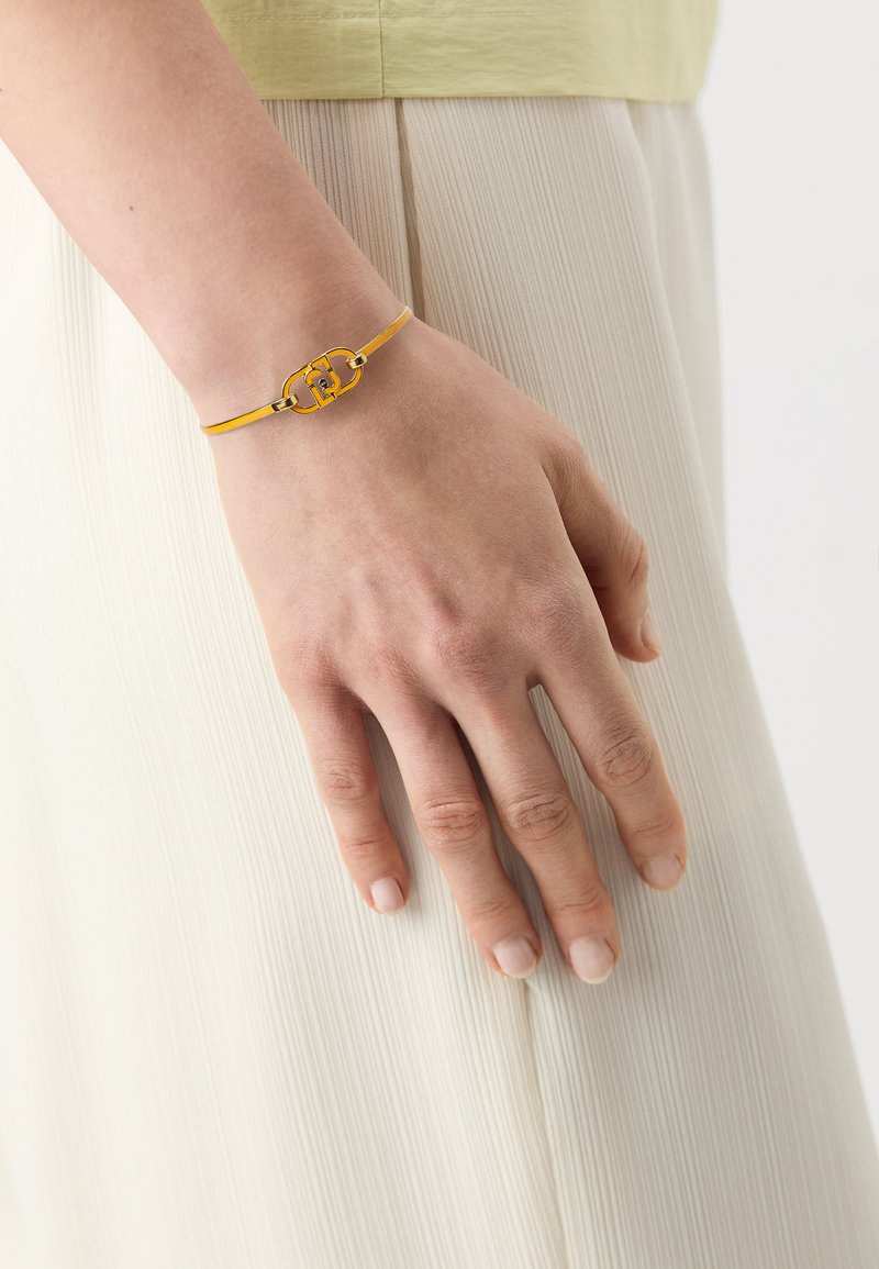 Gul bracelet med en metallisk låsdetalj, båret på en hånd med naturlige negler, mot en dunklet, lysfarget fargebakgrunn.