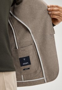 Innenraum eines grau-braunen Wollblazers mit einem dunkelblauen Markenschild, Etikett für schmale Passform und weißer Paspelierung entlang der Kanten. Eine einzelne Tasche ist sichtbar.