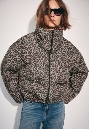 Žena nosiaca slnečné okuliare, zimnú bundu s leopardím vzorom zapnutú na zips a modré rifle, stojaca s rukami vreckách proti jednoliatemu pozadiu.