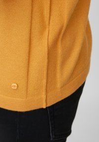 Pull tricoté en jaune vibrant avec un ourlet côtelé et un panneau latéral. Présente un bouton décoratif près de l'ourlet. Tissu doux et texturé.
