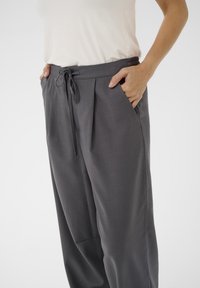 Pantalon gris plissé avec taille à cordon de serrage et poches latérales, présentant une texture de tissu lisse et une coupe décontractée.