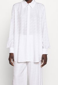 Persona di carnagione scura che indossa una camicia bianca a maniche lunghe e larga, abbottonata, insieme a pantaloni larghi coordinati, su uno sfondo chiaro e semplice.