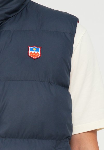 Navy blue padded vest with high collar and red embroidered shield logo featuring blue and yellow accents. Smooth texture and minimalist design.