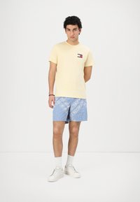 Gul kortermet t-skjorte med en kontrastlogo, kombinert med blå mønstrede badebukser. Hvite sneakers og strømper fullfører antrekket.