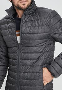 Giacca trapuntata grigia con collo alto, chiusura a zip e tasche laterali. Il tessuto ha una finitura texture. Design leggero.