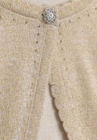 Goldfaden gestrickte Strickjacke mit gewelltem Rand und einem dekorativen silbernen Blütenknopf, der den Verschluss akzentuiert. Weiche Textur.