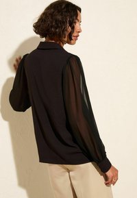 Blouse noire avec des manches bouffantes transparentes et longues, et un col. Corps en tissu doux texturé avec un détail froncé aux poignets. Associée à un pantalon beige.
