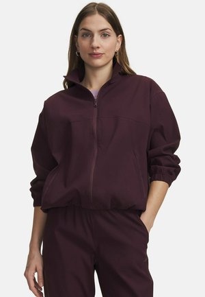 Vrouw draagt een donkerpaarse jas met ritssluiting, elastische manchetten en bijpassende broek, staand met één hand in de zak tegen een witte achtergrond.