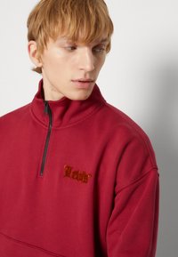 Felpa rossa con collo alto e zip a metà, con logo "Levi's" ricamato in marrone caldo, realizzata in materiale morbido e dal fitting rilassato.