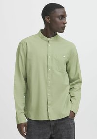 Camicia a maniche lunghe verde chiaro con colletto alla mandarino, tasca frontale e chiusura con bottoni. Il tessuto sembra morbido con una finitura testurizzata.
