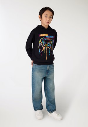 Ragazzo in piedi con le mani dietro la schiena, indossa una felpa nera con cappuccio e una grafica colorata di un controller di gioco, con jeans blu su uno sfondo bianco semplice.
