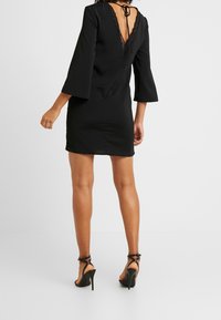 Robe noire avec un dos en V, ornée de dentelle et de manches évasées. Stylée avec des chaussures à talons moyens, soulignant une silhouette ajustée.