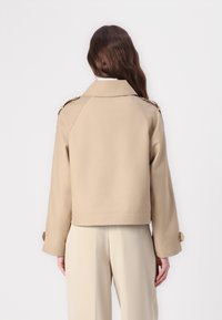 Giacca cropped beige con colletto a punta, maniche a raglan e dettagli con bottoni sui polsini. Tessuto liscio con un design minimalista.