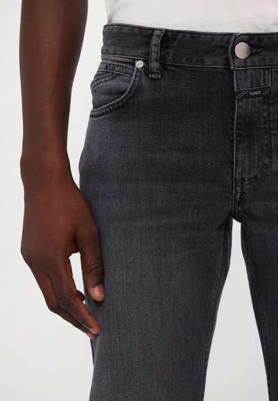Los jeans de mezclilla de color gris oscuro tienen un corte ajustado, costuras visibles, bolsillos frontales y un cierre de botón. El material tiene una ligera textura.