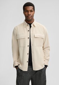 Beige corduroy shirt with two chest pockets, button front, and a collared neckline. Worn over a black top, paired with dark patterned pants.