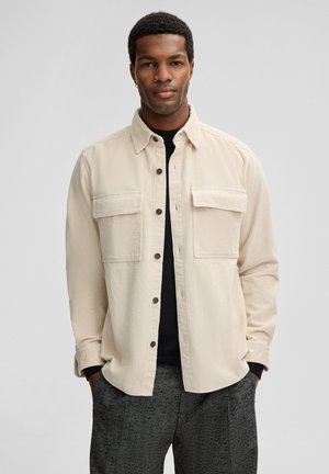Beige corduroy shirt with two chest pockets, button front, and a collared neckline. Worn over a black top, paired with dark patterned pants.