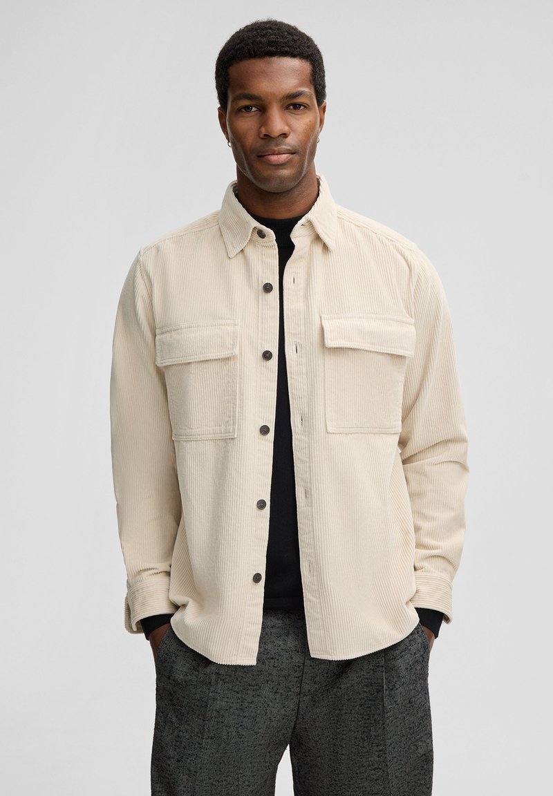 Beige corduroy shirt with two chest pockets, button front, and a collared neckline. Worn over a black top, paired with dark patterned pants.