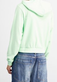 Světle zelená mikina s kapucí na šňůrky, žebrovanými manžetami a lemem. Dokončeno s modrými džínovými kraťasy s velkými zadními kapsami.