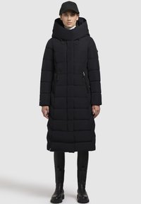 Lunga giacca a vento nera con un colletto alto, trapuntatura verticale, due tasche laterali con zip e un cappuccio. Indossata sopra pantaloni neri aderenti e stivali.
