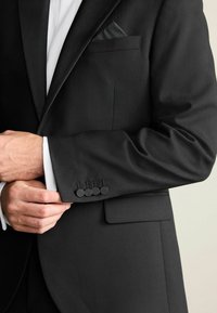 Sacou negru cu o textură netedă, rever clasic și o borsetă. Manșetele au patru butoane negre, iar dedesubt se află o cămașă albă.
