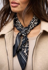 Černý a béžový hedvábný šátek s leopardím vzorem a pruhovanými akcenty, elegantně uvázaný kolem krku, v kombinaci s béžovým topem a sakem.