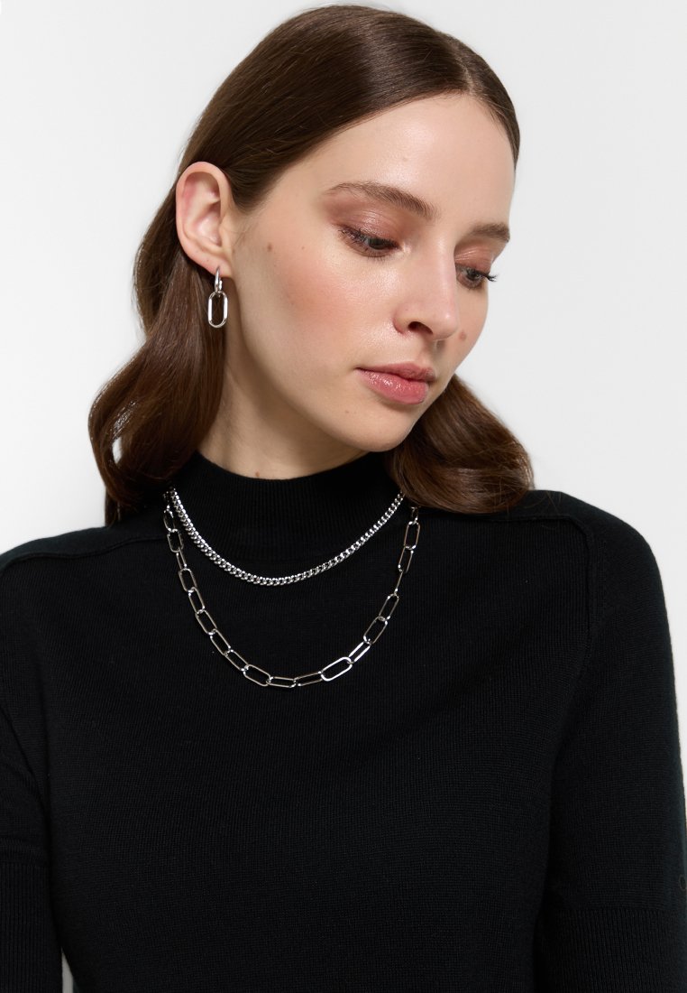 Silberne Schmuckstücke aus Metall, bestehend aus einer massiven Kettenhalskette und einer zarten Kette, getragen über einem schwarzen hochgeschnittenen Pullover.