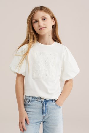 Ragazza con lunghi capelli biondi che indossa una maglietta bianca con maniche a sbuffo e dettagli in pizzo e jeans azzurri, in piedi con una mano in tasca.