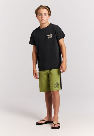 Black short-sleeve T-shirt with logo on the chest, paired with green swim shorts featuring a black accent and graphic print. Black flip-flops.