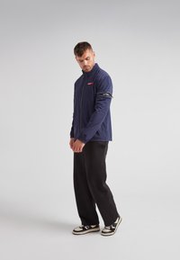 Chaqueta ligera azul marino con un alto cuello, cierre de cremallera y logotipo rojo. Combinada con pantalones de chándal negros y zapatillas en blanco y negro.