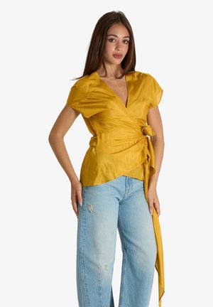 Giovane donna in piedi che indossa una camicetta avvolgente color senape a maniche corte e jeans azzurri strappati su sfondo bianco.