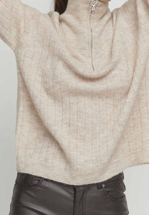 Femme portant un pull en maille côtelée beige avec une demi-fermeture éclair et un pantalon en cuir noir, les bras levés au-dessus de la tête.