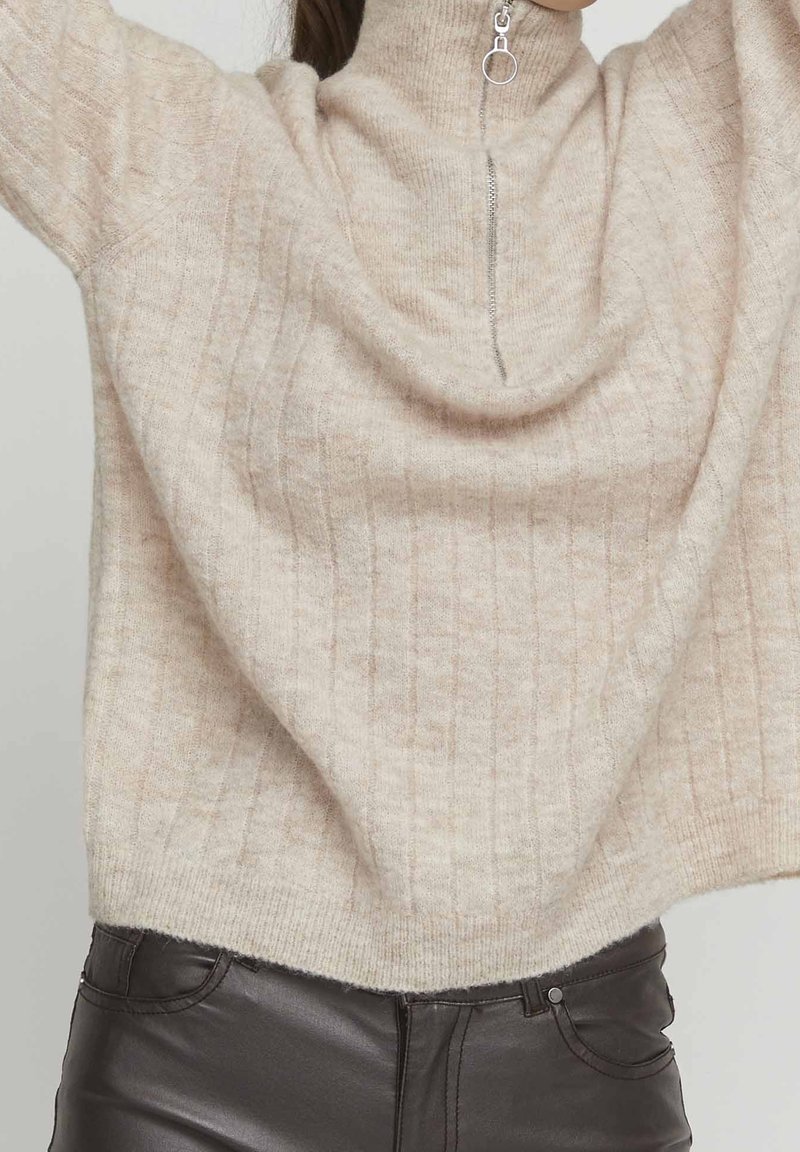 Femme portant un pull en maille côtelée beige avec une demi-fermeture éclair et un pantalon en cuir noir, les bras levés au-dessus de la tête.