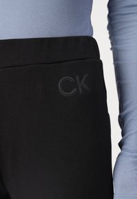 Crne rastezljive hlače glatke teksture, s toniranim "CK" logom na pojasu, u kombinaciji s prilagođenim svijetloplavim majicom dugih rukava.