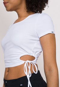 Top corto bianco a costine con maniche corte, dotato di aperture laterali e dettagli con cordino per una vestibilità regolabile. Dettaglio del logo nero sulla manica.