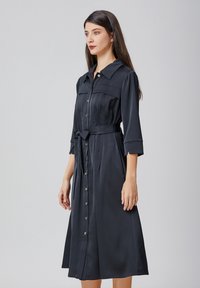 Robe noire à manches longues avec un col, manches trois-quarts et taille ceinturée. Texture lisse et détails plissés.