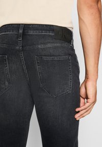 Jean en denim gris foncé avec une coupe slim, présentant un patch en cuir de la marque sur la ceinture et deux poches arrière aux bords incurvés.