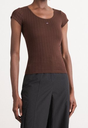 Vrouw draagt een nauwsluitend bruin geribd kortermig shirt en zwarte, hooggetailleerde broek, staand tegen een witte achtergrond.