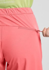 Pantaloni sportivi di un vivace colore rosa fucsia con una vita testurizzata e una tasca con cerniera sul retro, con una maglia verde a contrasto visibile sopra.