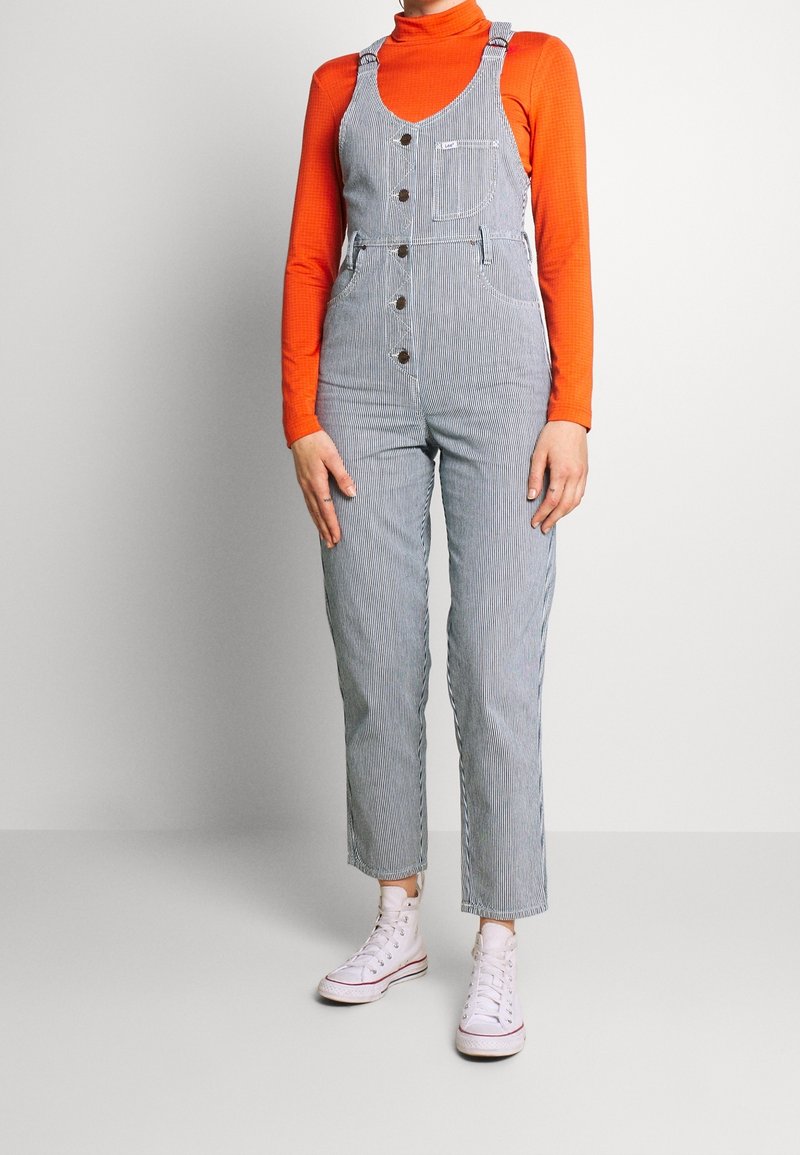 Salopette en denim rayée avec un corsage ajusté, de larges bretelles, des poches avant et des boutons marron, associée à un col roulé orange et des baskets blanches.
