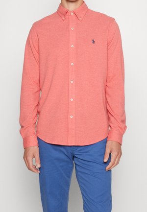 Homme portant une chemise corail à boutons avec un logo de cheval bleu et un pantalon bleu, debout les bras détendus le long du corps sur un fond uni.