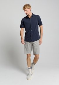 Navy short-sleeve button-up shirt paired with light gray shorts; model wears white sneakers with navy and gray striped socks.