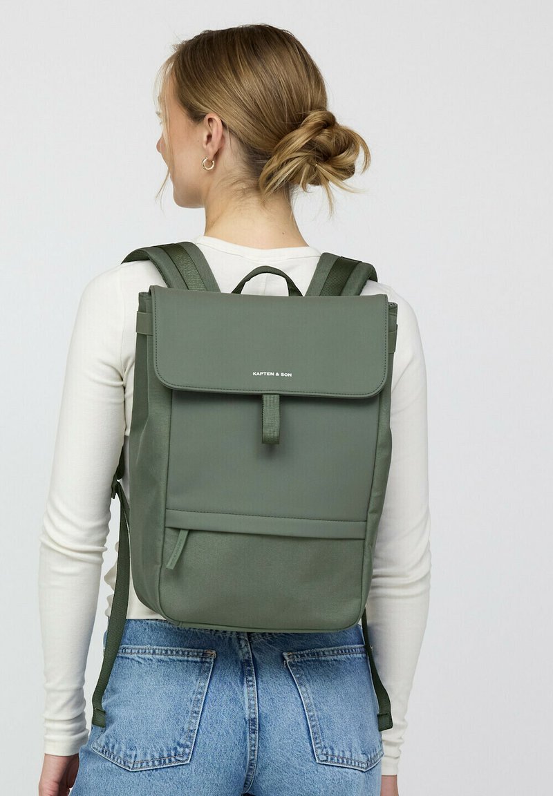 Groene canvas rugzak met een klepdesign, voorzak, verstelbare banden en minimalistische logodetail. Gladde textuur en rechthoekige vorm.