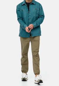 Giacca di jeans teal con quattro tasche sul petto, abbinata a pantaloni color sabbia. Le calzature presentano un mix di colori bianco e mimetico con suola testurizzata.