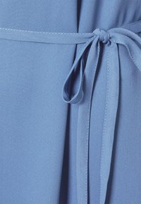 Světle modrá tkanina se hladkým texturem; obsahuje detail zavázaného pasu a kontrastní švy podél okrajů.