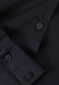 Must mustriga, mis on valmistatud siledast ja kergest kangast, pika varrukaga, klassikalise krae ja kahe musta nööbiga mansetil.