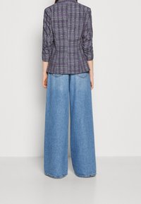 Veste à carreaux en violet foncé et vert, cintrée à la taille avec des manches trois quarts, associée à un pantalon en denim bleu clair à jambe large.