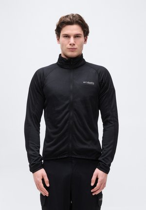 Moški v črni Columbia Titanium jakni z zadrgo stoji pred enobarvnim belim ozadjem.