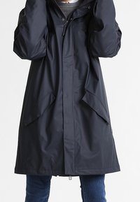 Czarny wodoodporny płaszcz z kapturem, wyposażony w dwie duże przednie kieszenie, zapięcie na zatrzaski i zamek oraz długie luźne rękawy.