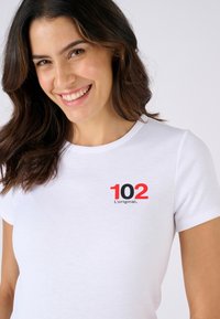 T-shirt en coton blanc avec un col rond et des manches courtes, portant sur la poitrine le texte en noir et rouge "102 L'original."